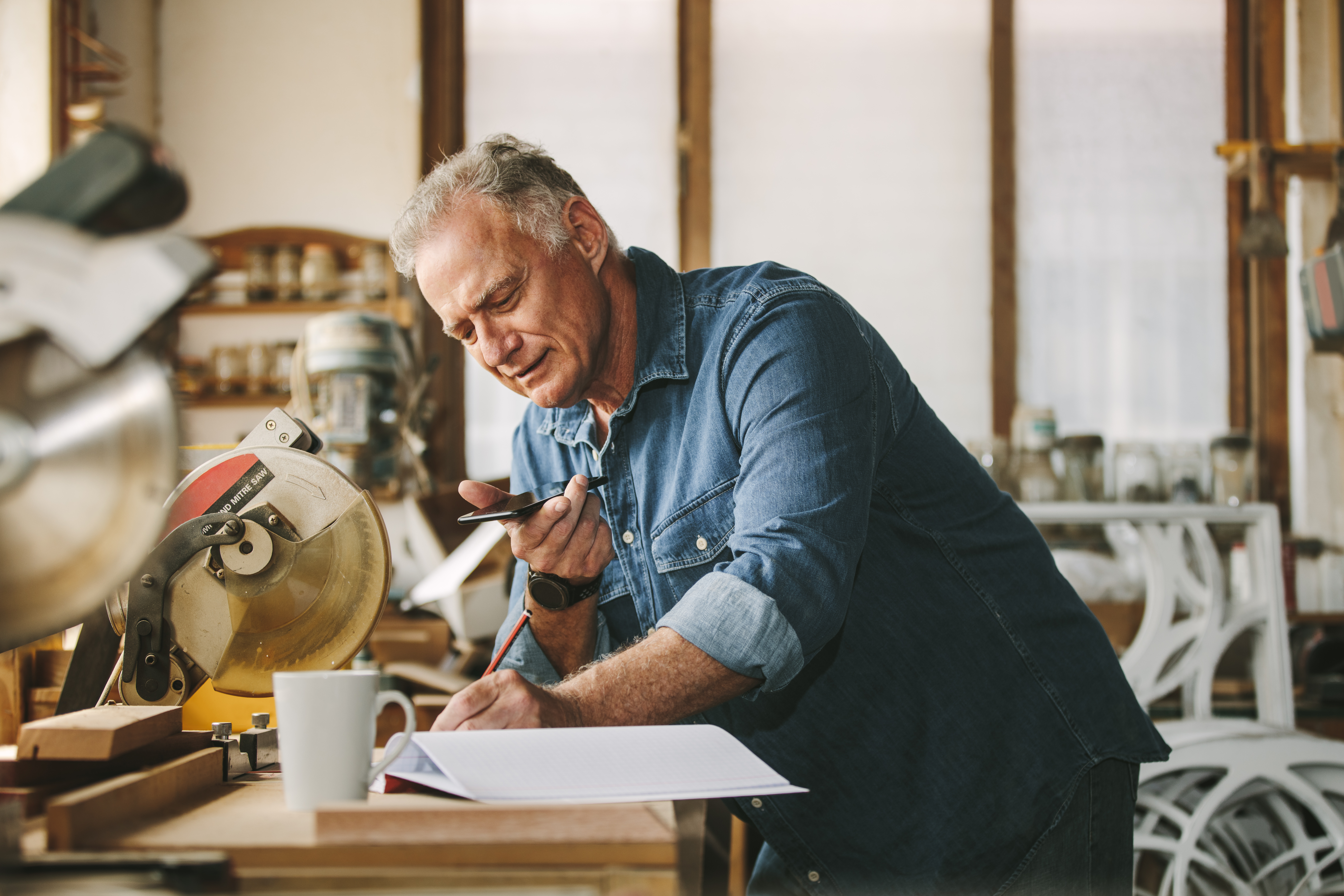 Senior carpenter working at his workshop while standing at desk using mobile phone and writing in a book. Mature man working in carpentry workshop.