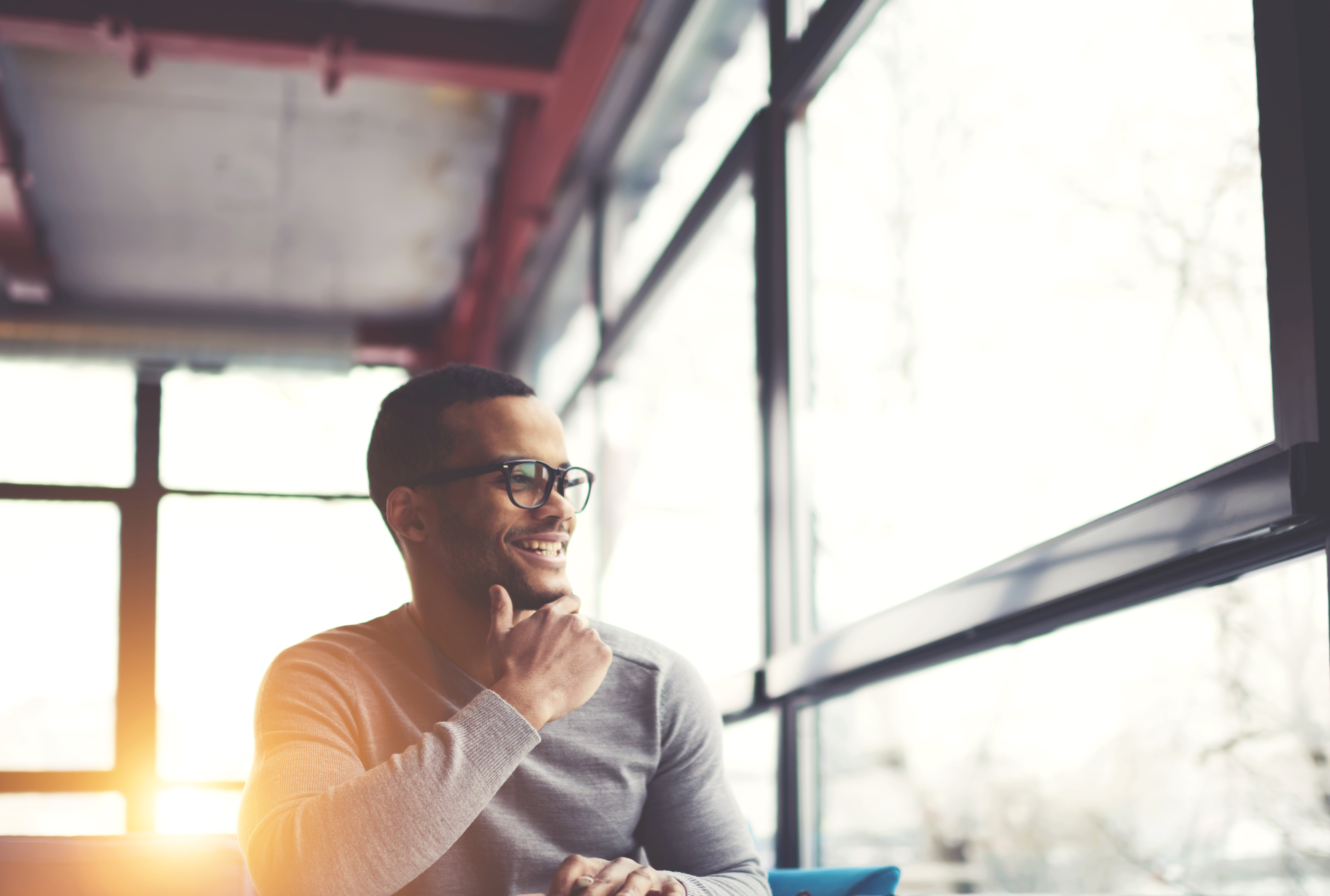 Cheerful afro american hipster guy in trendy spectacles sitting in loft interior cafe resting in good mood, smiling young dark skinned businessman satisfied with successfully completed project