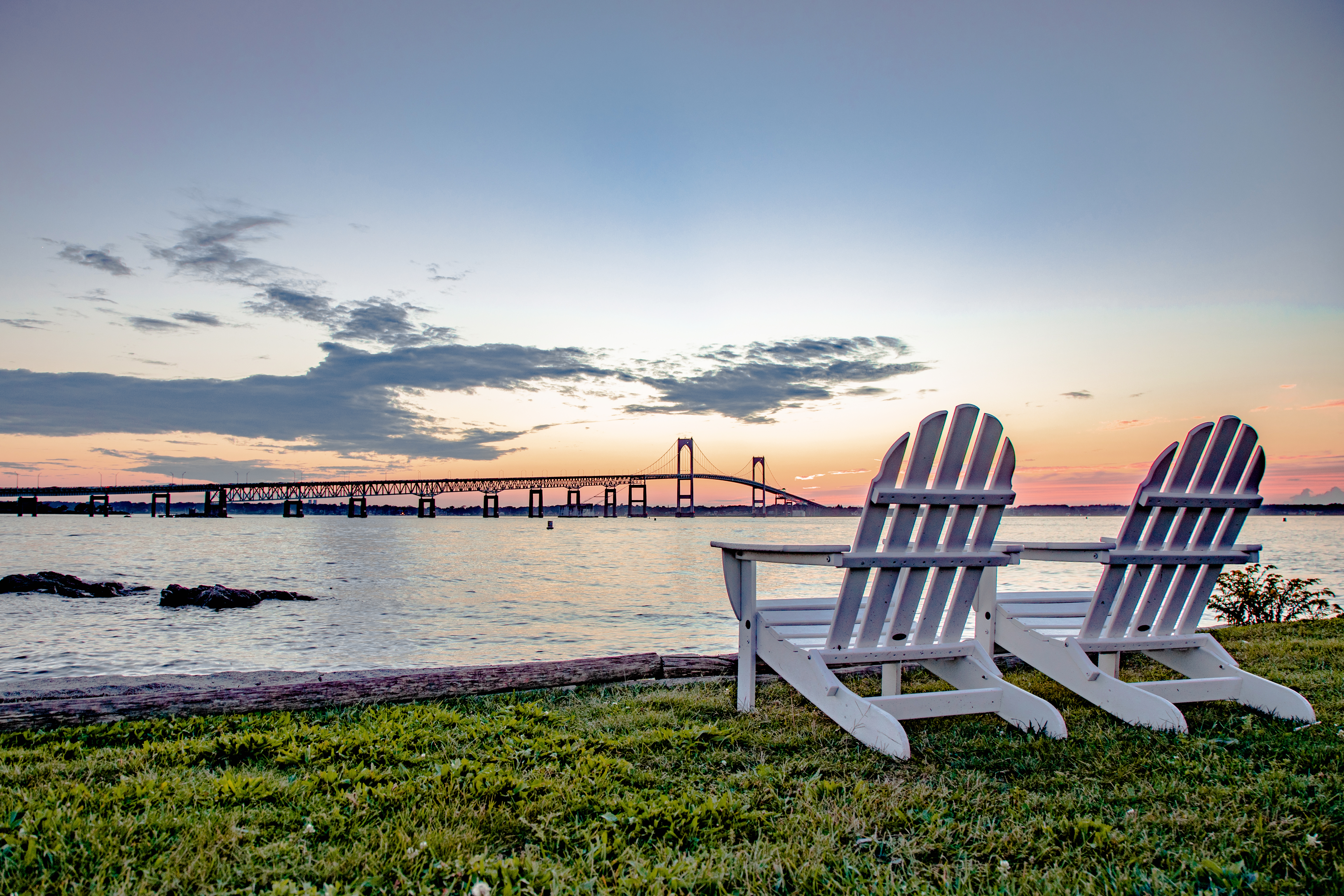 Pair of Adirondack chairs on the edge of the water with a large suspension bridge in the background.