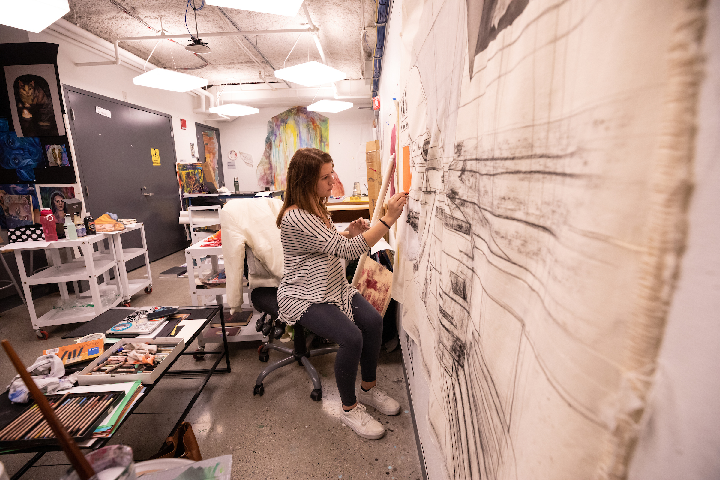 Image 2 of 5: Female student sitting and drawing on paper taped to the wall